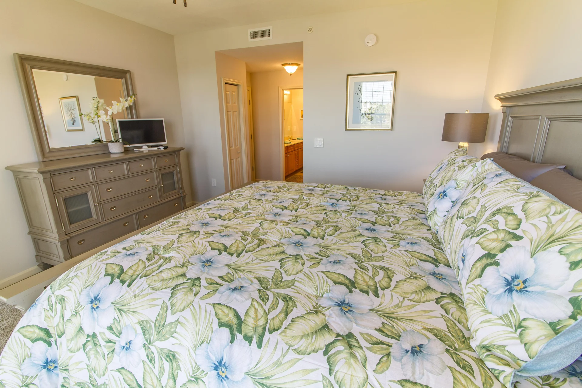 King bed with floral bedding faces a taupe wood dresser topped by a large mirror and a small flat-screen TV.