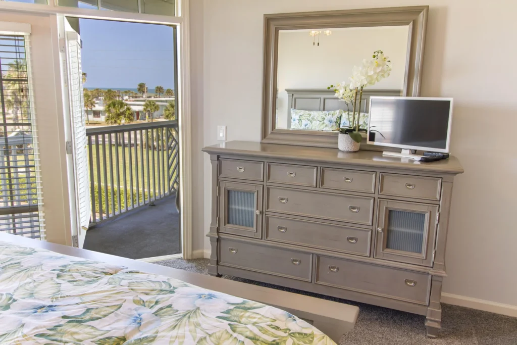 Gray wooden dresser with mirror, small flat-screen TV, and orchid beside the open balcony door in the bedroom.