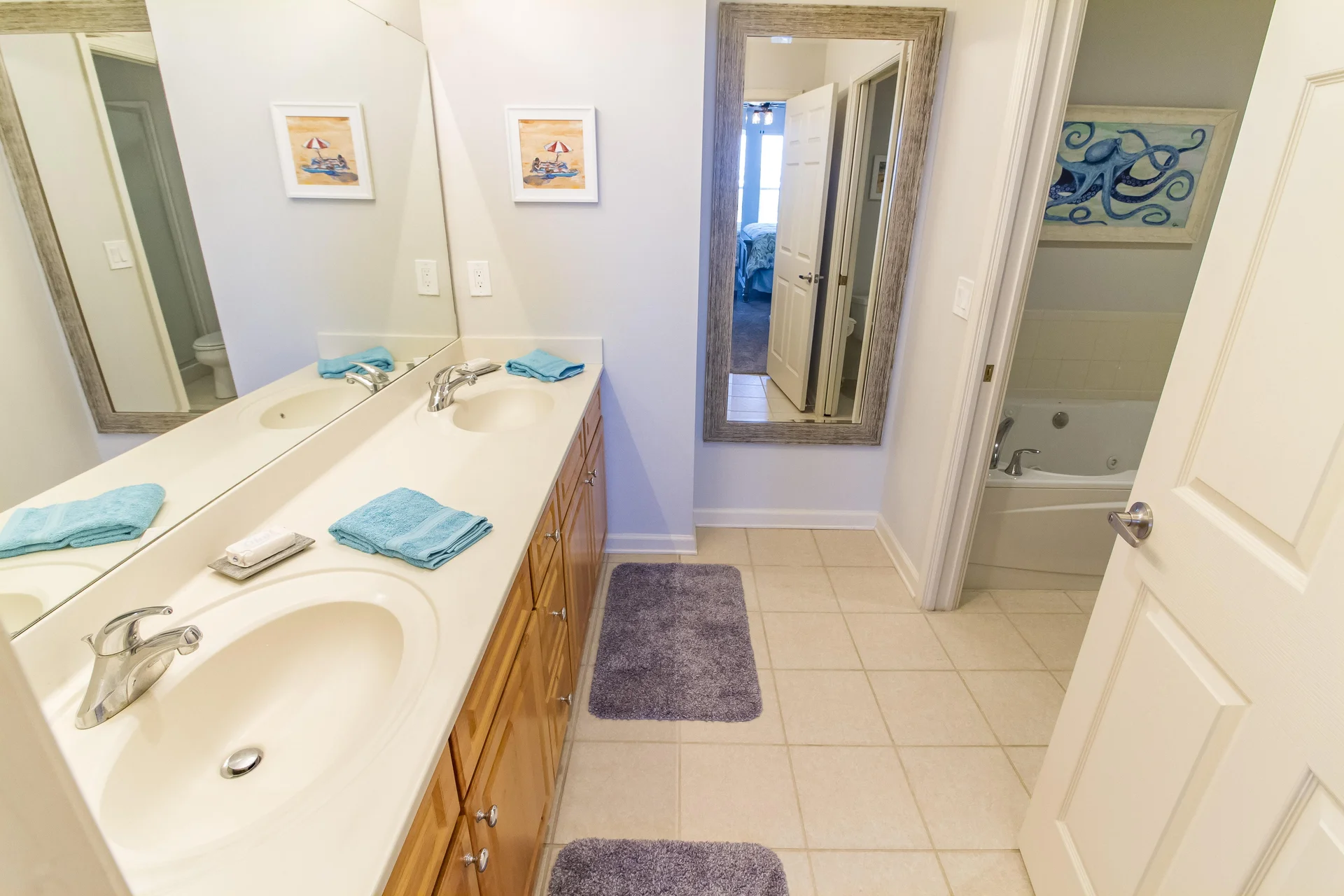Bathroom vanity with dual sinks, light wood cabinets, large mirror, blue hand towels, and tiled floor mats.