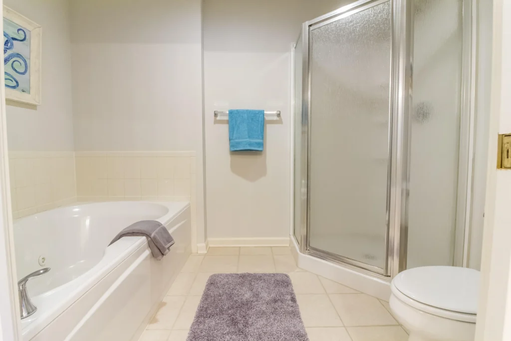 Bathroom with corner glass shower, white soaking tub with gray towel, beige tiled floor, and blue wall art.