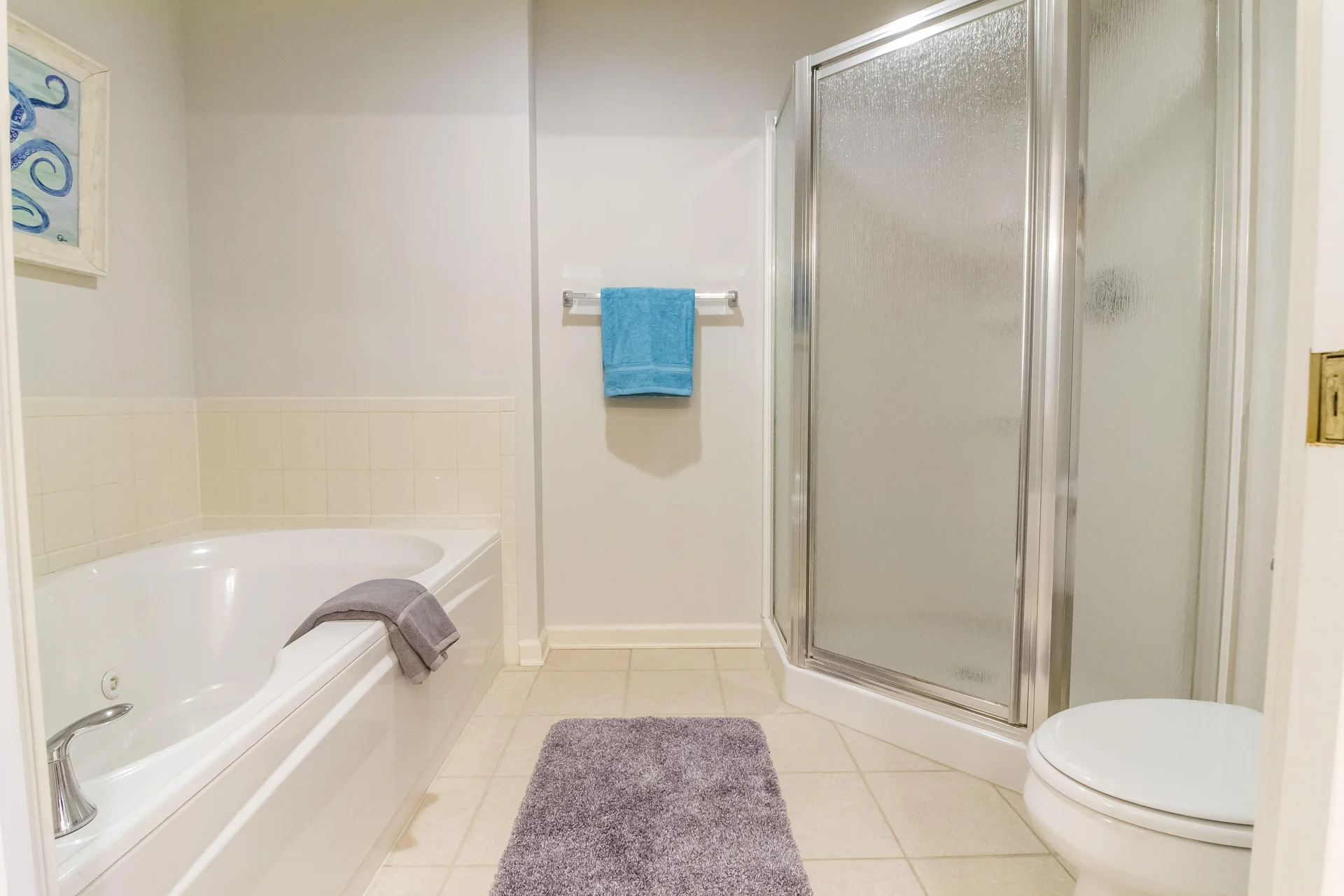 Bathroom with corner glass shower, white soaking tub with gray towel, beige tiled floor, and blue wall art.