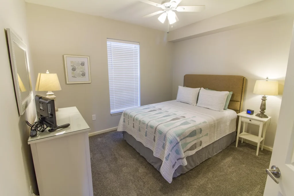 Neutral-toned bedroom features a beige upholstered headboard, patterned quilt, white nightstand with lamp, and carpeted flooring.