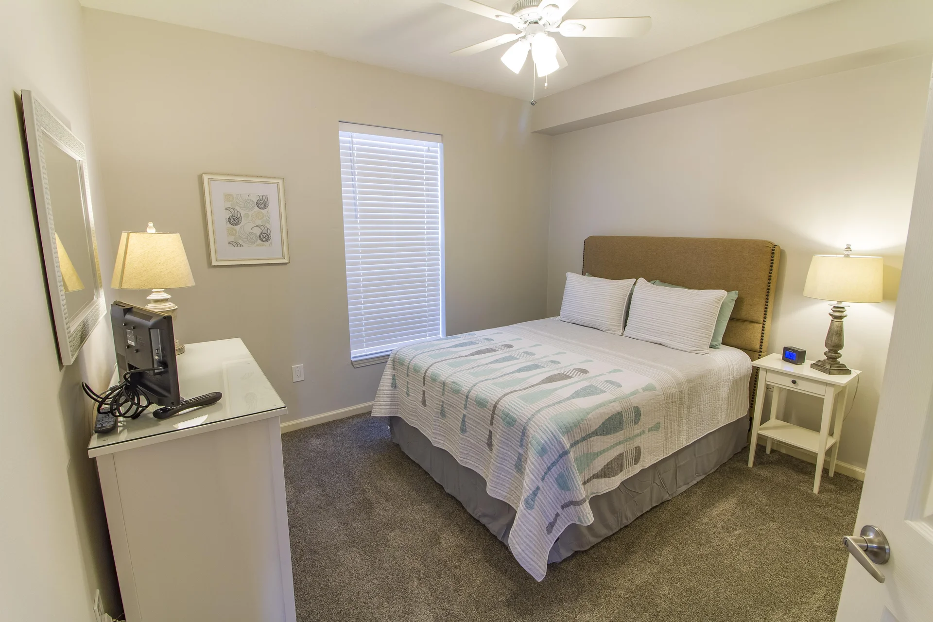 Neutral-toned bedroom features a beige upholstered headboard, patterned quilt, white nightstand with lamp, and carpeted flooring.