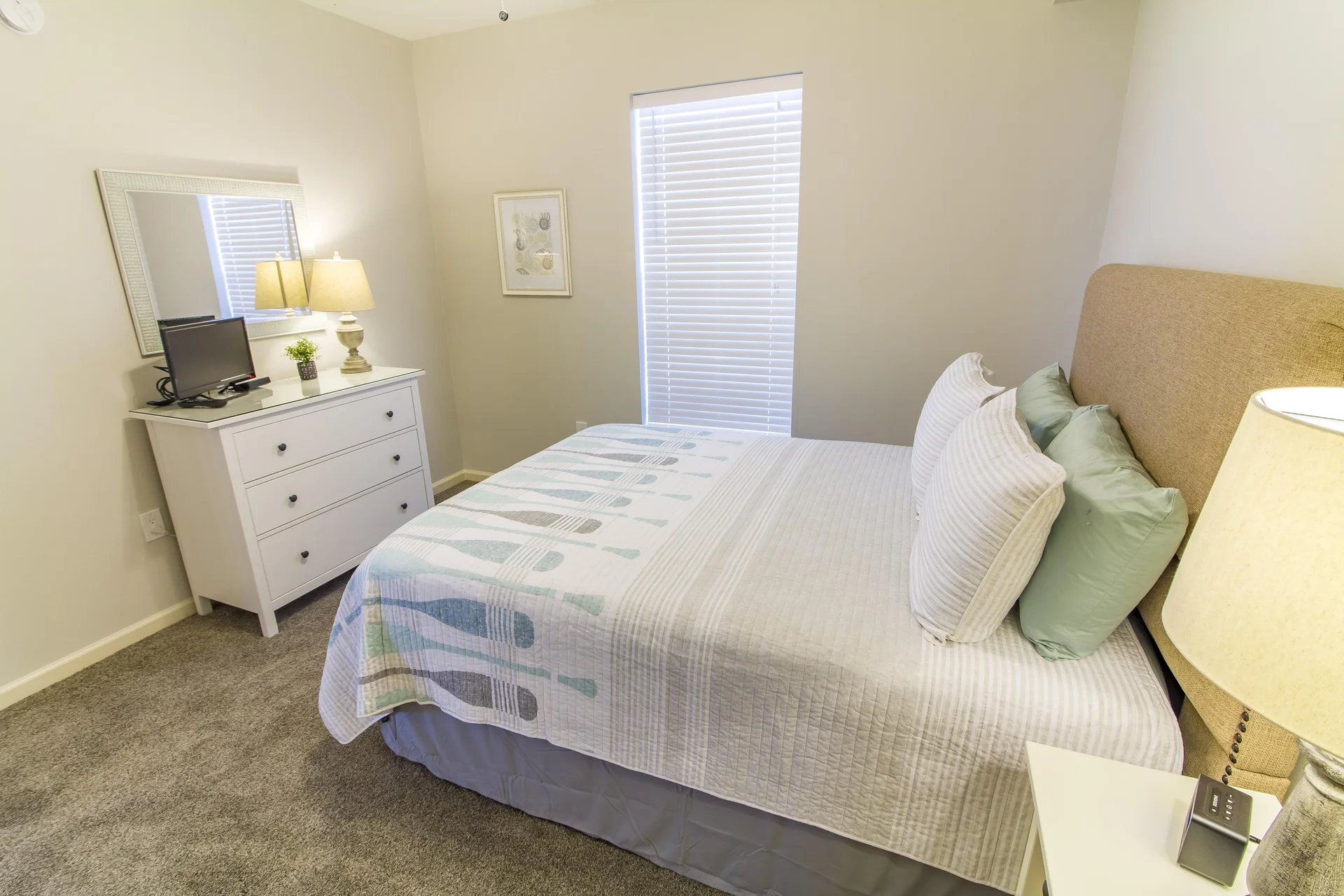 Bed with patterned quilt and layered pillows positioned beside a beige upholstered headboard and a nightstand lamp.