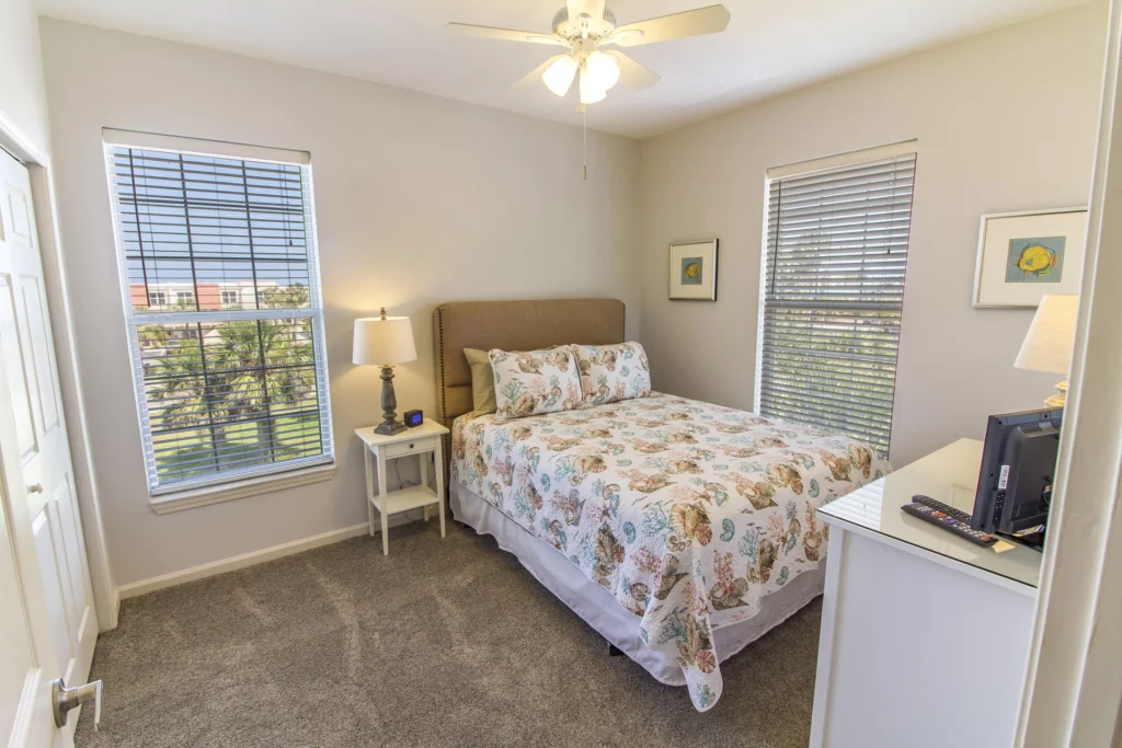 Carpeted bedroom with twin windows featuring white blinds, a white nightstand with lamp, and ceiling fan above.