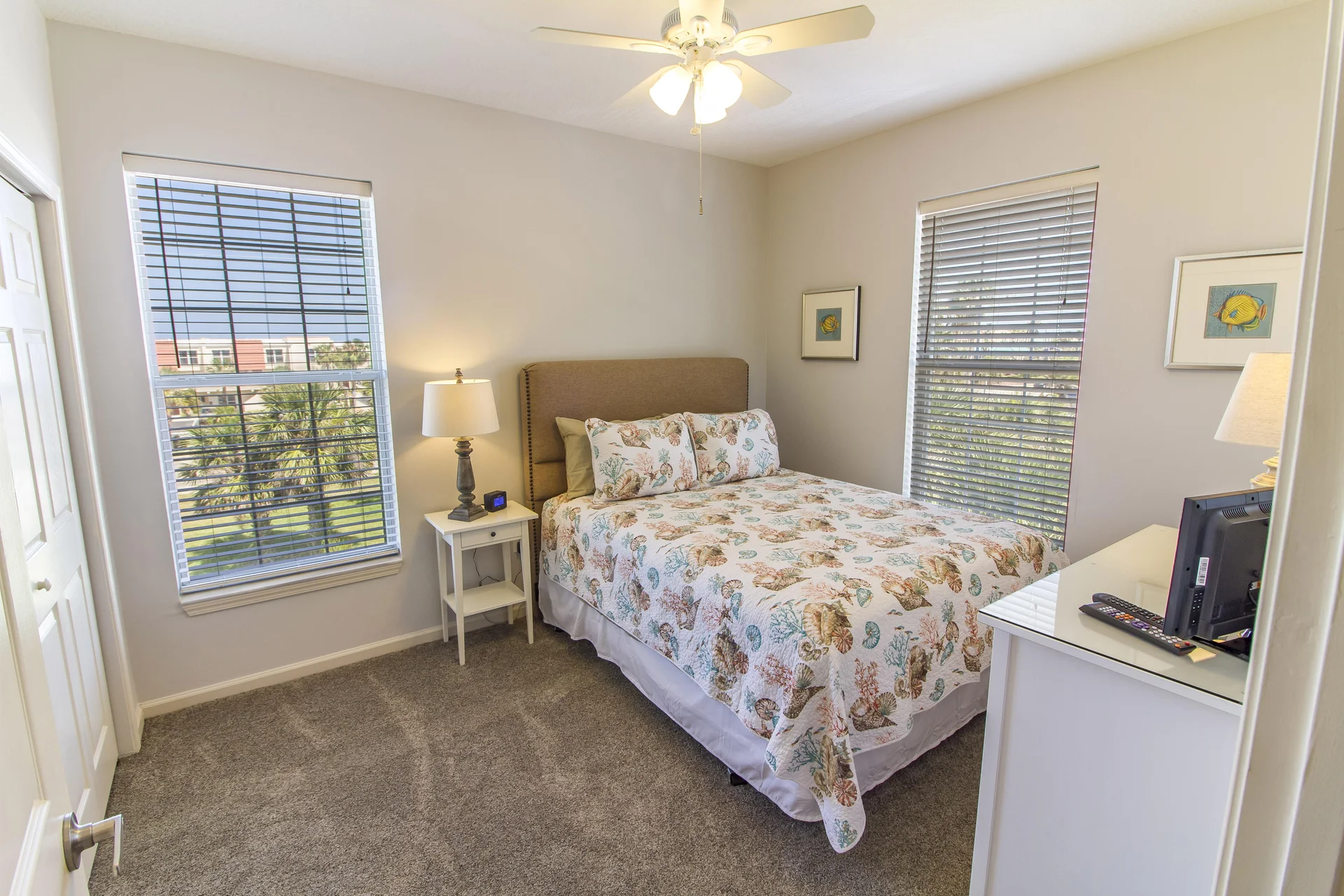 Carpeted bedroom with twin windows featuring white blinds, a white nightstand with lamp, and ceiling fan above.
