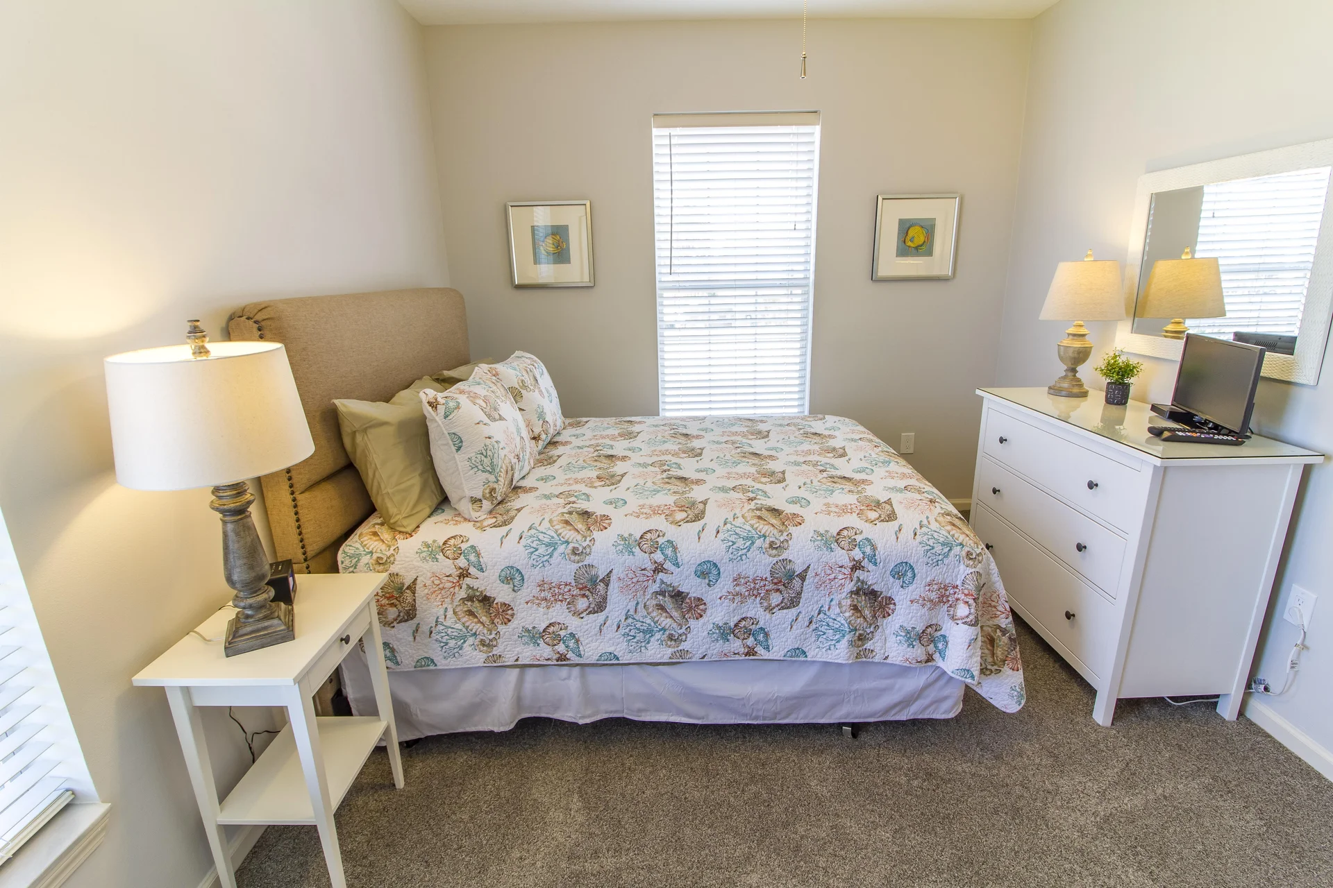 Window with white blinds sits between two framed seashell prints above the queen bed with shell-patterned quilt.
