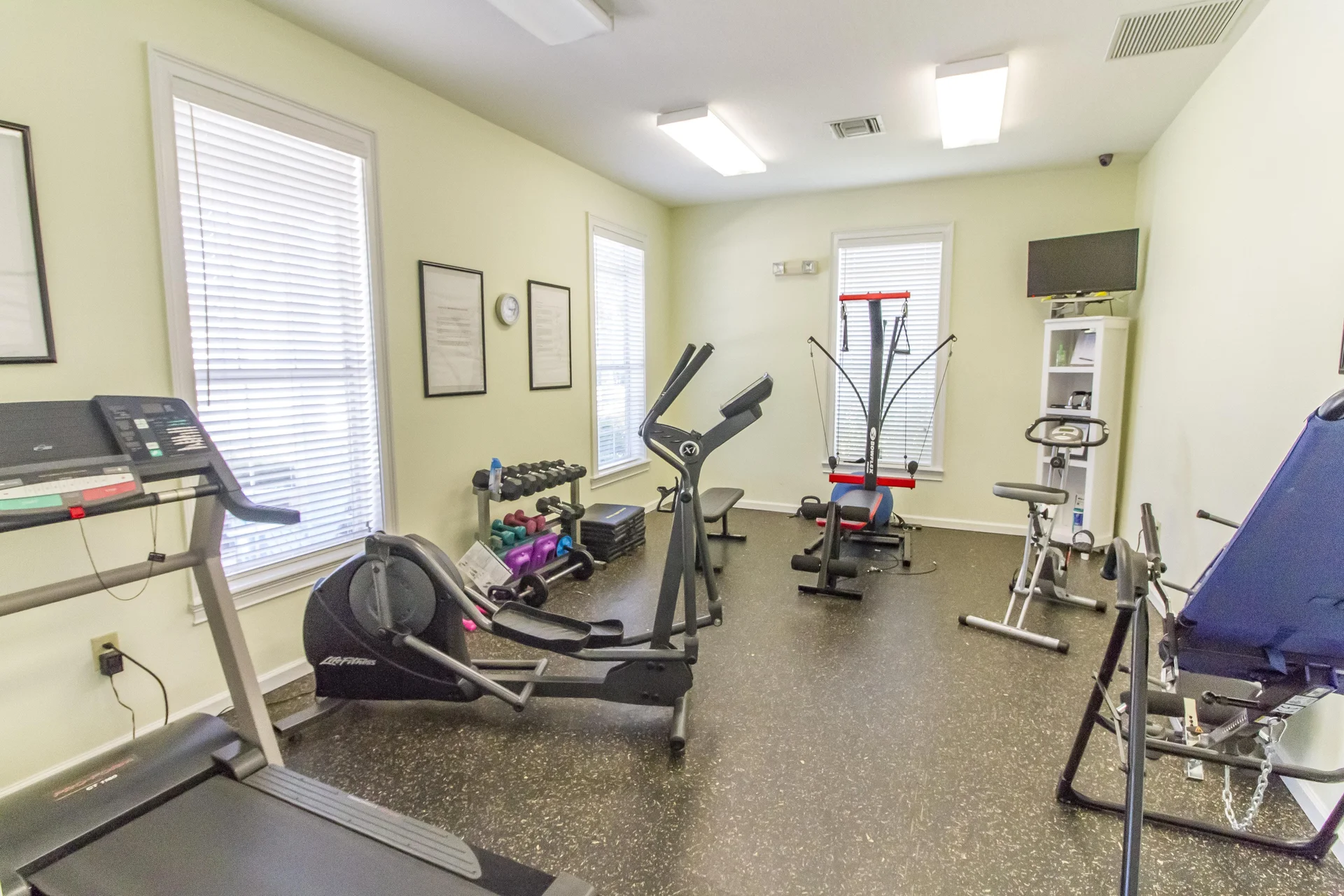 Fitness room with treadmill, elliptical, weight bench, stationary bike, dumbbells, and wall-mounted TV.