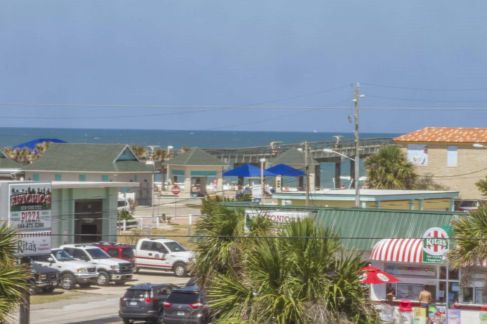 Partial ocean view beyond palm trees, parked vehicles, and local shops with colorful awnings and signs.