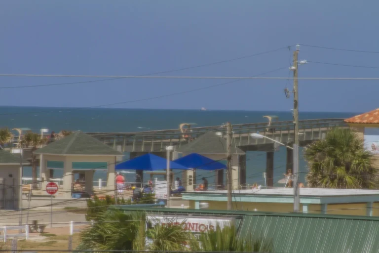 View of the ocean and pier framed by palm trees, blue umbrellas, and rooftops under a clear sky.