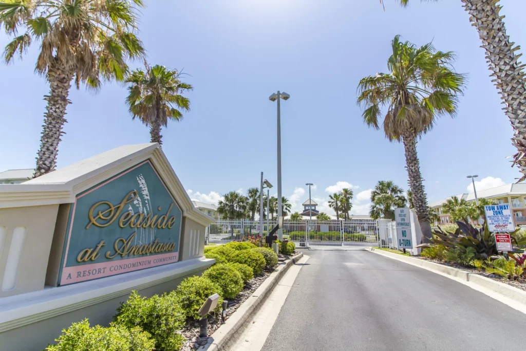 Entrance gate to Seaside at Anastasia resort with palm trees, landscaped shrubs, and clear blue sky overhead.