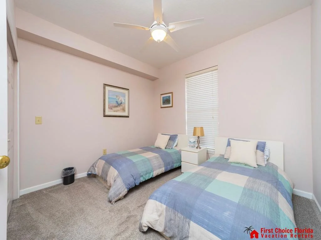 Two twin beds with pastel patchwork quilts flank a white nightstand with a gold lamp beneath a window with blinds.