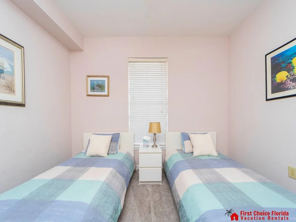 Two twin beds with pastel patchwork quilts flank a white nightstand topped with a brass lamp.