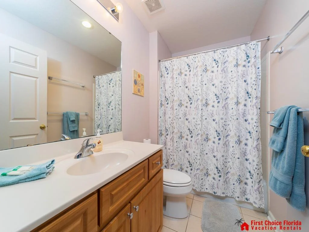 Single-sink vanity with honey wood cabinets and a large mirror overlooks a tiled bathroom with floral shower curtain.