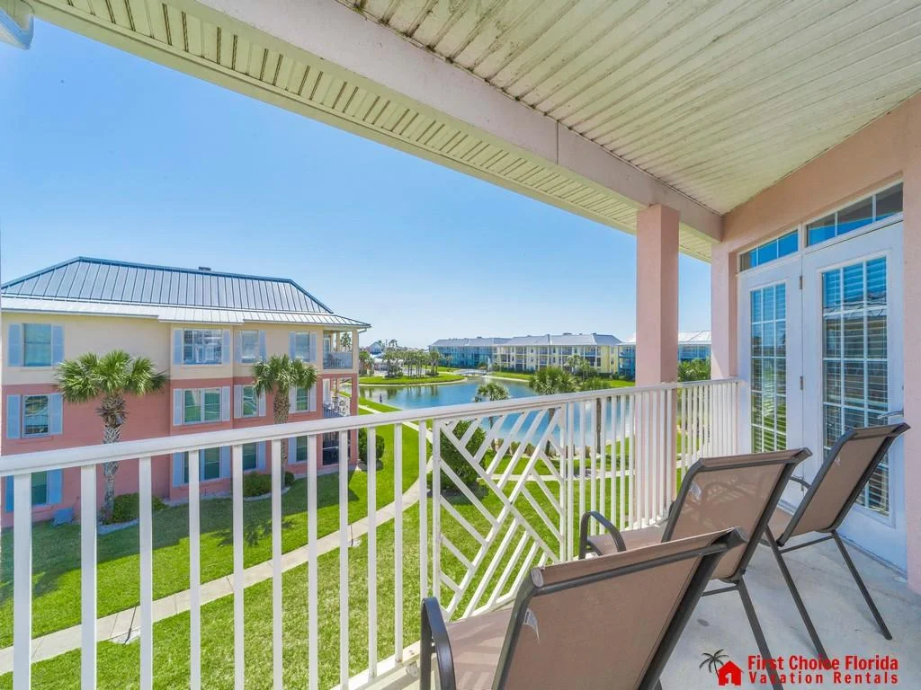 Covered balcony with white railing overlooks manicured lawns, palm trees, and a tranquil pond with pastel condo buildings.