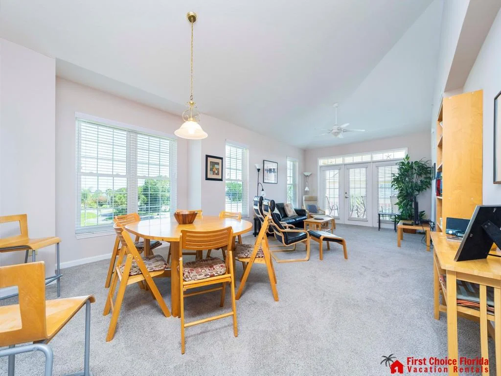 Light gray carpeted living room features honey wood furniture, black leather chairs, and large windows with white blinds.
