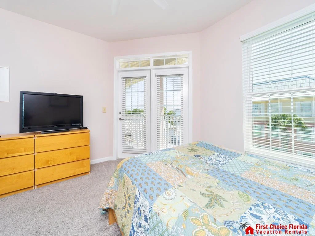 Light gray carpet frames a bed with a pastel floral patchwork quilt beside a dresser and TV.