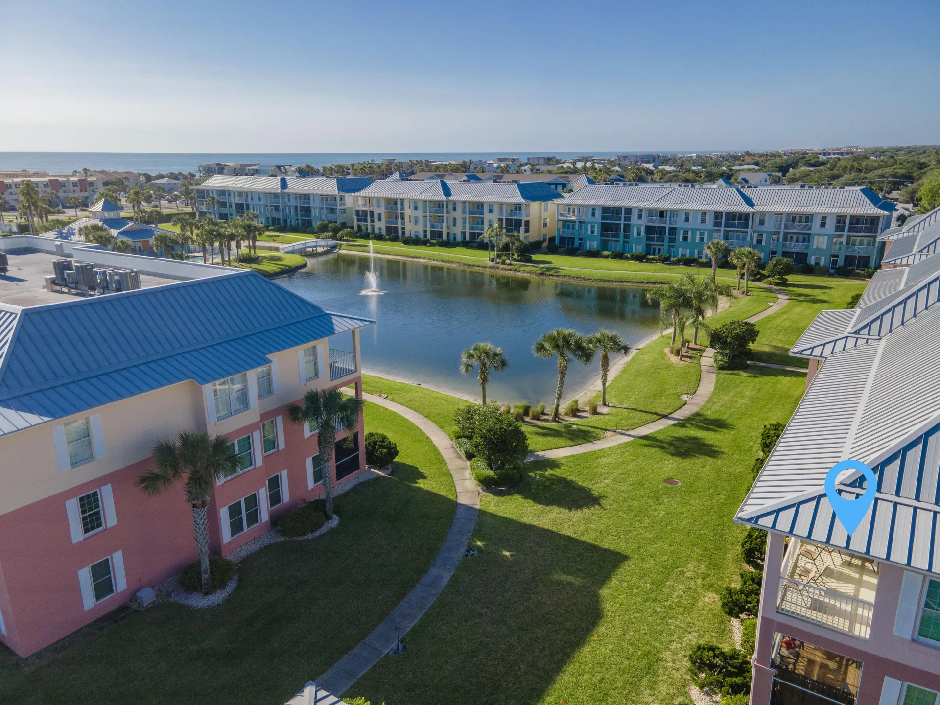 Aerial view of pastel-colored condo buildings surrounding a pond with a central fountain and palm-lined walking paths.