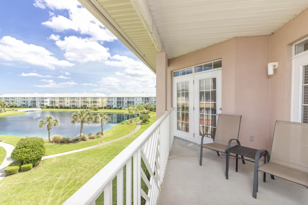 Balcony with beige mesh chairs and small table overlooks pond, palm trees, and pastel-colored condos.