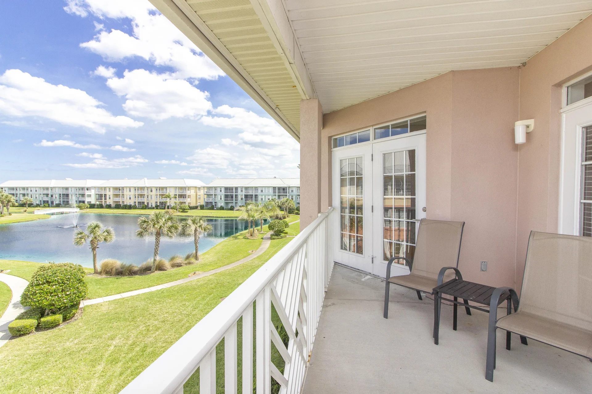 Balcony with beige mesh chairs and small table overlooks pond, palm trees, and pastel-colored condos.