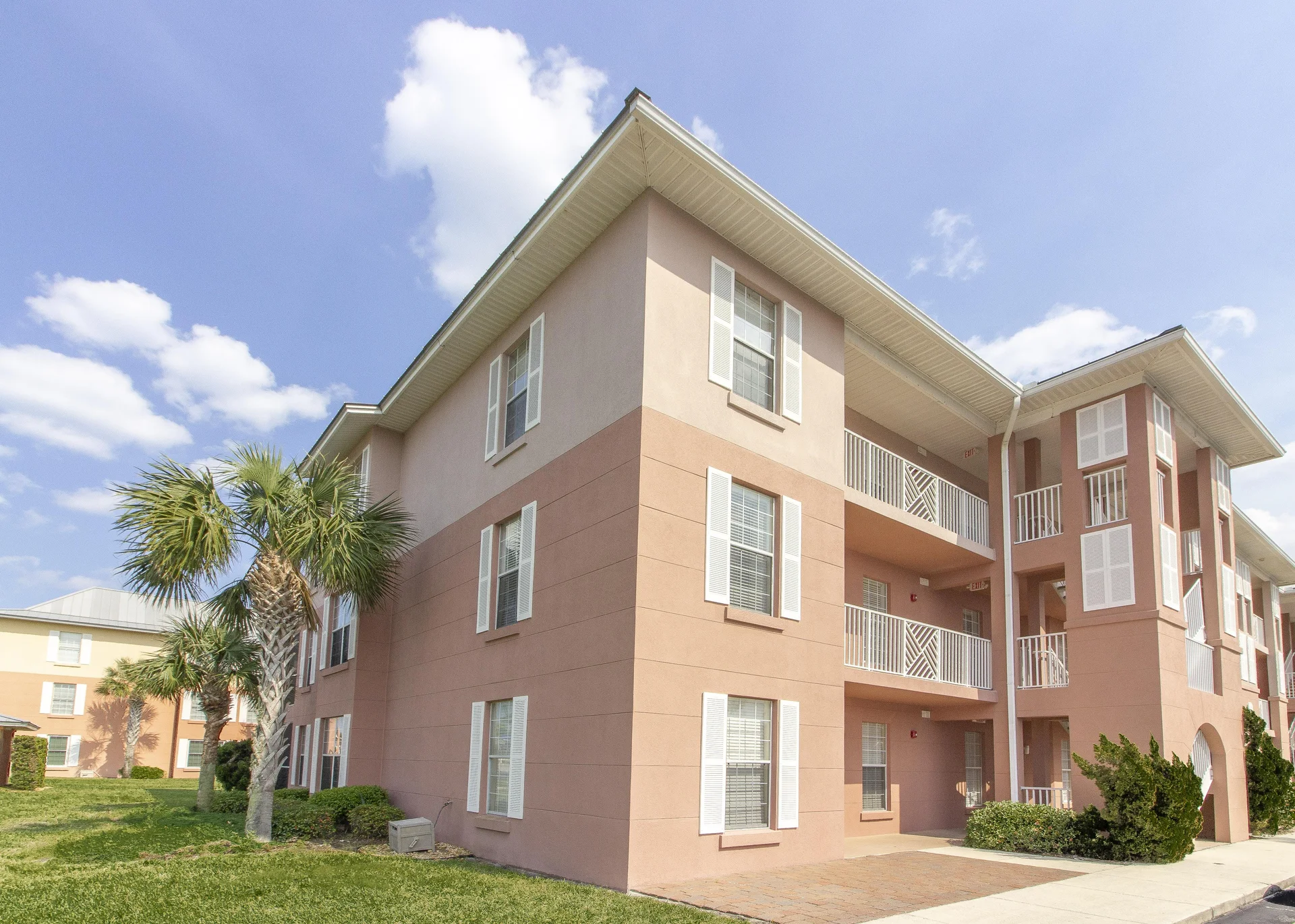 Exterior view of a three-story condo building with peach and tan stucco, white trim, and palm trees on a sunny day.
