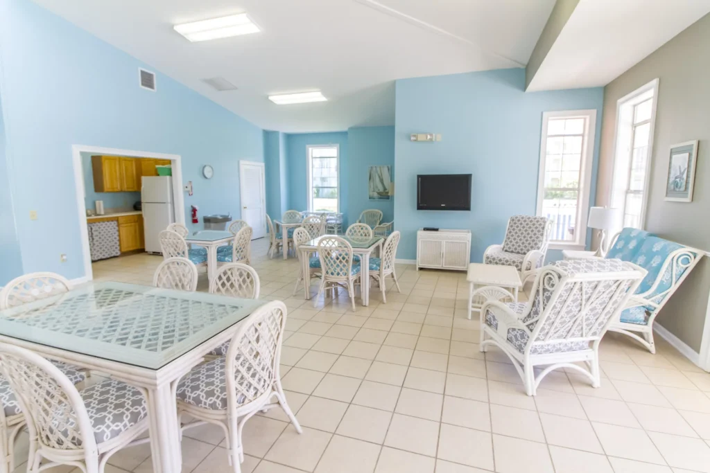 Community lounge with white wicker chairs, glass-top tables, blue walls, tiled floors, and a wall-mounted TV.