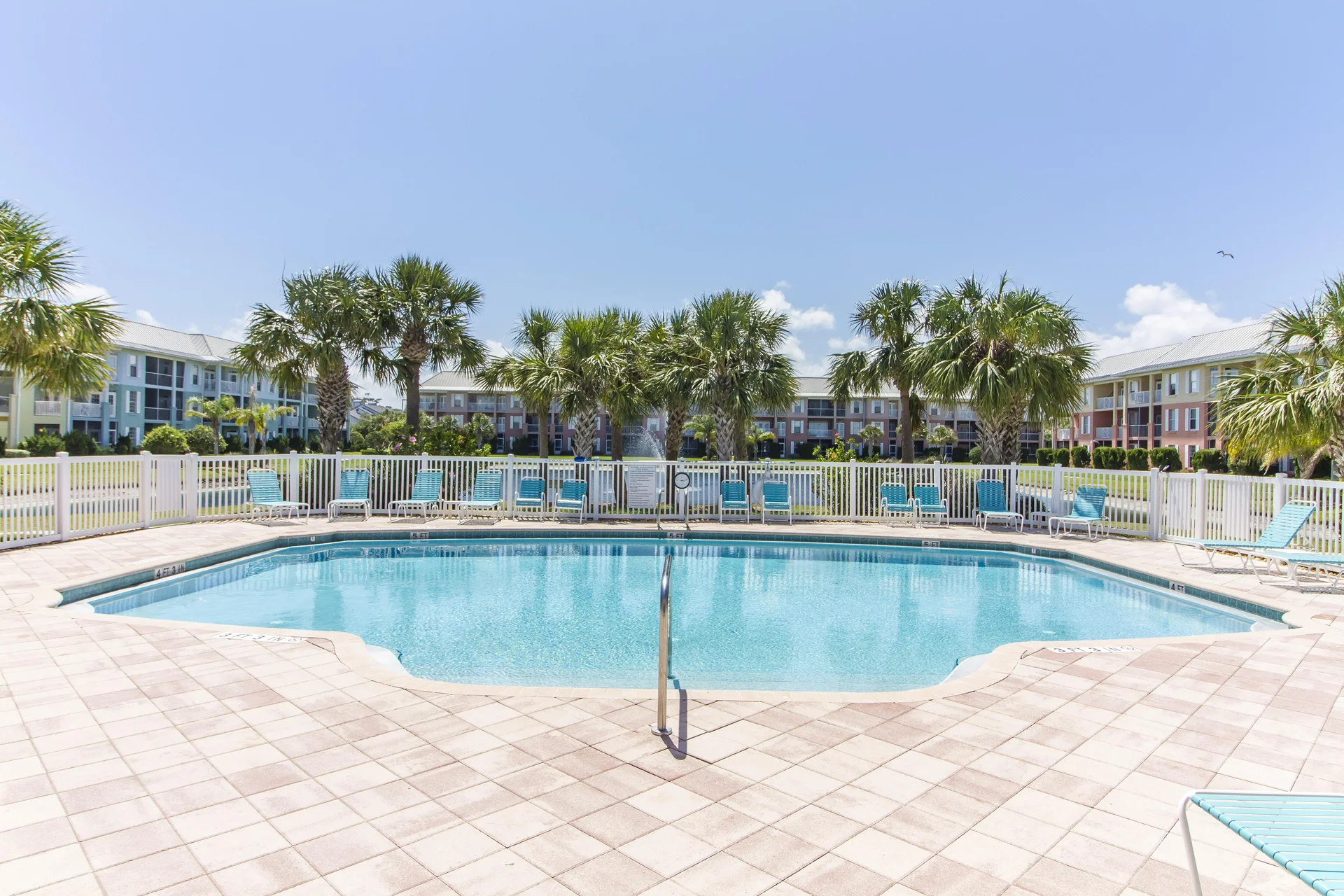 Outdoor pool with clear blue water, surrounded by tiled deck, white fence, palm trees, and colorful condo buildings.