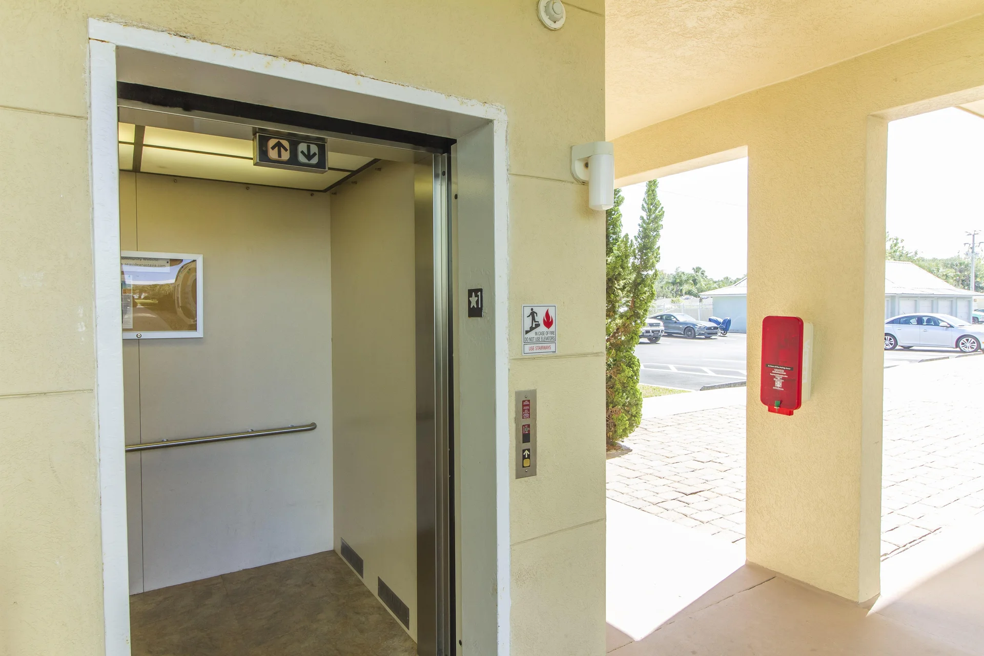 Elevator entrance with beige walls, stainless steel trim, and outdoor parking visible through open corridor.
