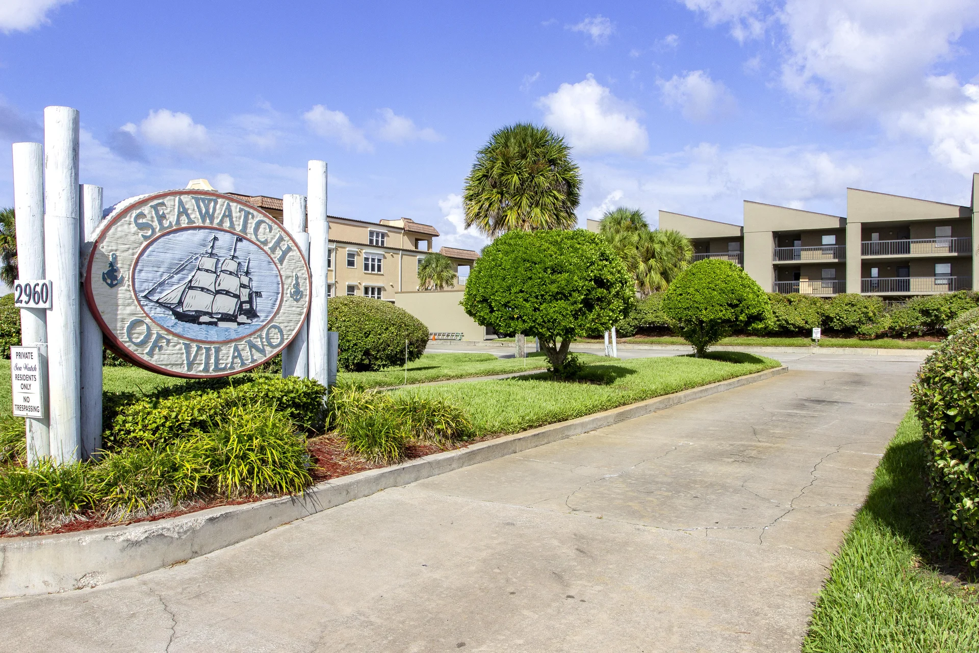 Concrete driveway leads into the Seawatch condo complex, bordered by manicured green shrubs and palm trees.