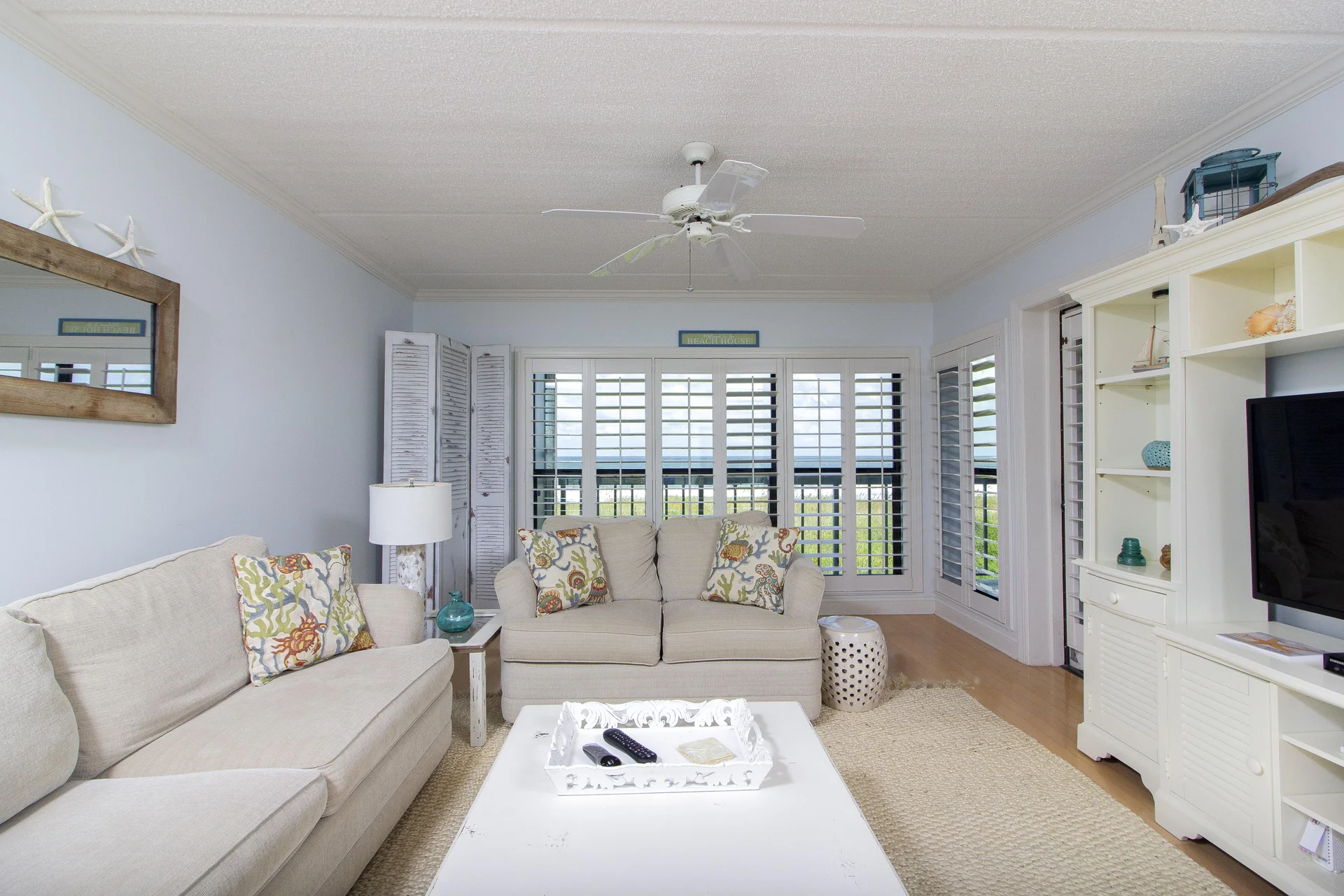 Living room with beige sofas, coastal-patterned pillows, white coffee table, and plantation shutters framing an ocean view.
