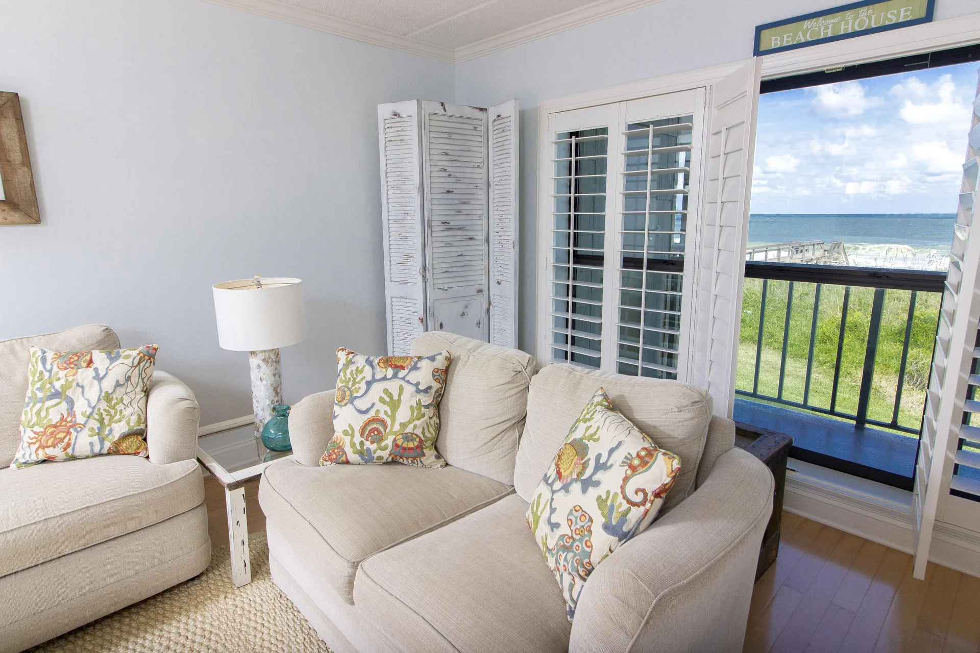 Second-floor living area with beige sofas, coral-patterned pillows, white distressed side table, and ocean views through plantation shutters.