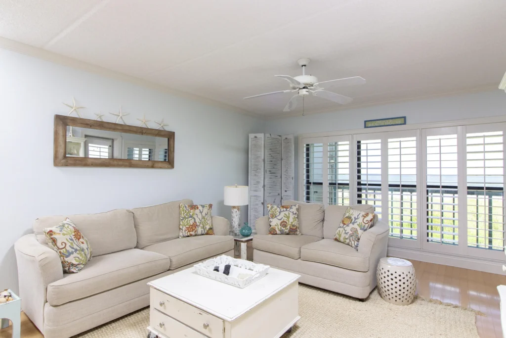 Living room with beige sofas, patterned pillows, white distressed coffee table, and plantation shutters overlooking the balcony.