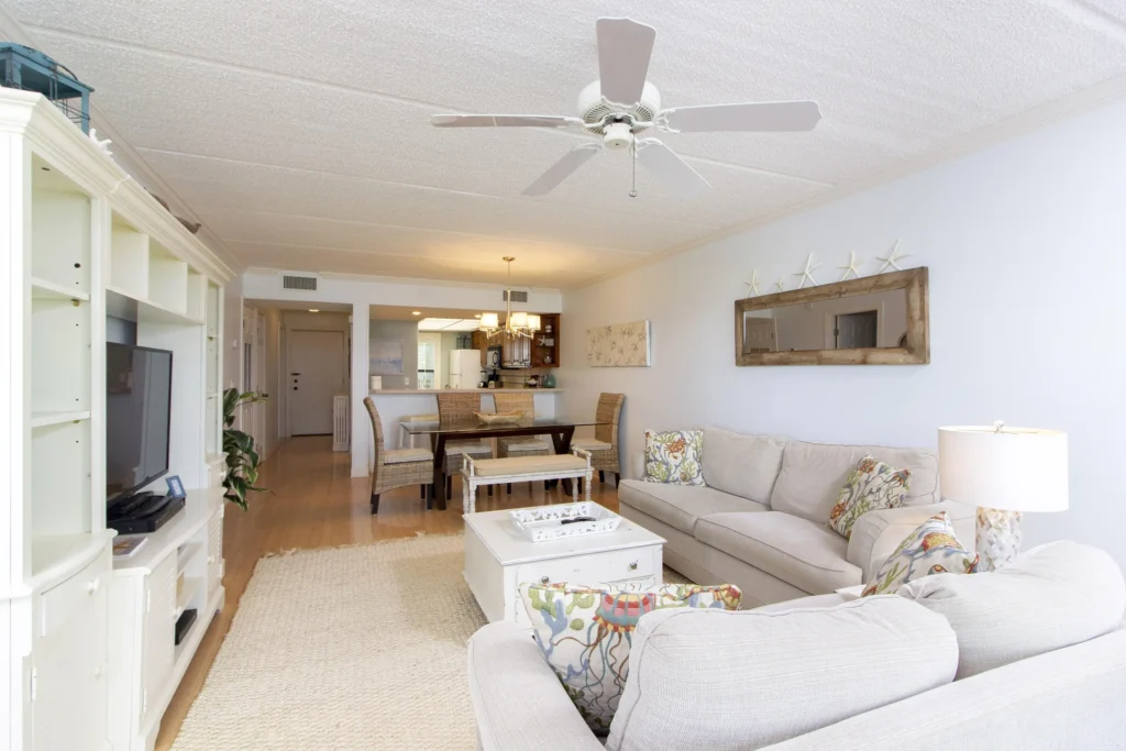 Open living and dining area with beige sofas, coastal-patterned pillows, white entertainment center, and hardwood floors.
