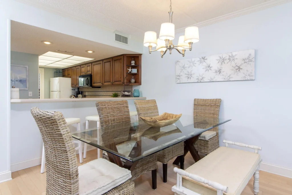 Dining area with glass-top table, woven chairs, cushioned bench, and brushed nickel chandelier overhead.