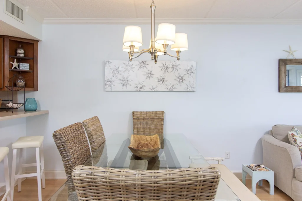 Glass-top dining table surrounded by woven rattan chairs beneath a brushed nickel chandelier.