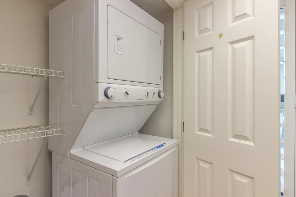 Stacked white washer and dryer sit next to white shelving inside a compact laundry closet with beige walls.
