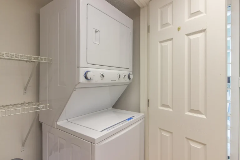 Stacked white washer and dryer sit next to white shelving inside a compact laundry closet with beige walls.
