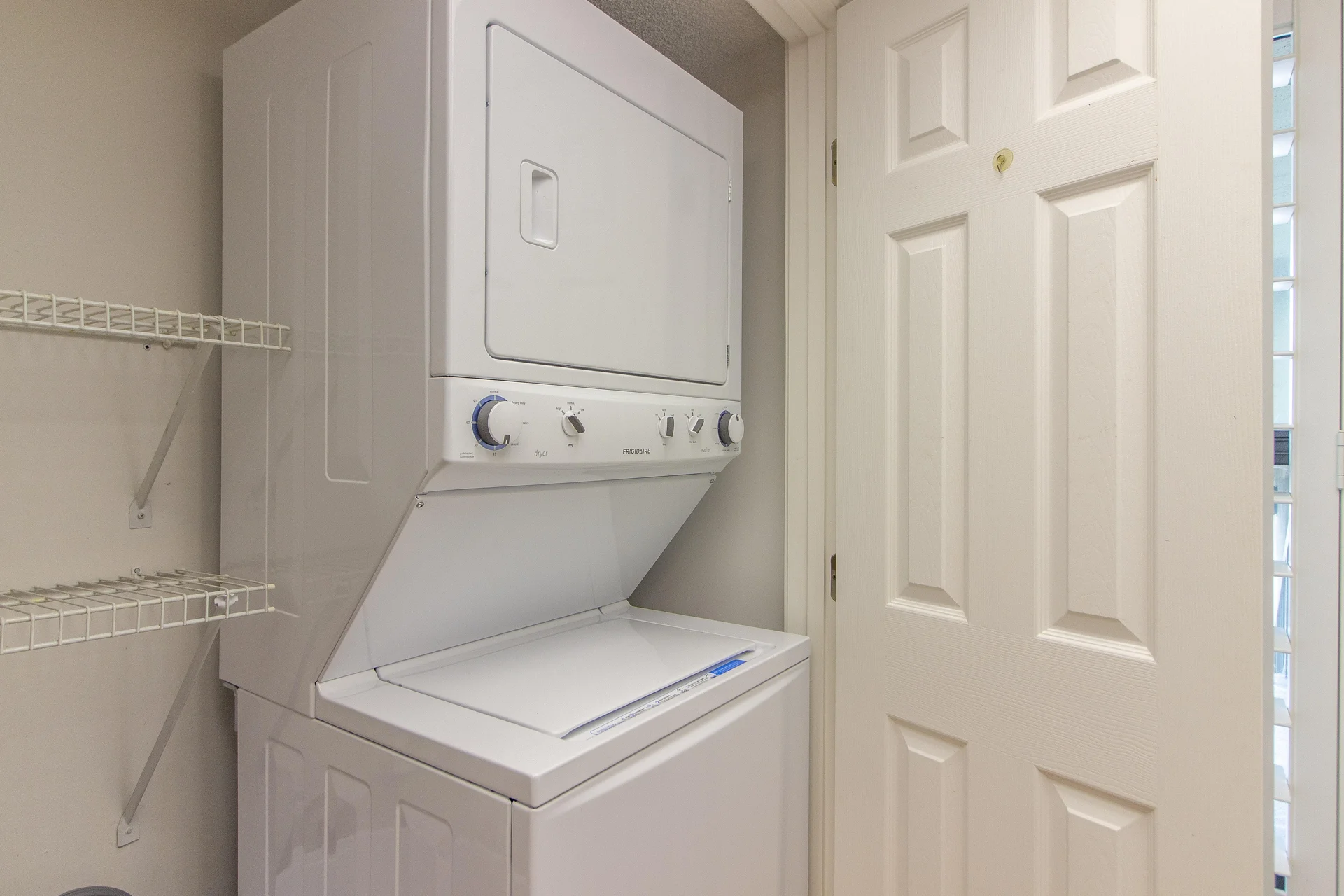 Stacked white washer and dryer sit next to white shelving inside a compact laundry closet with beige walls.