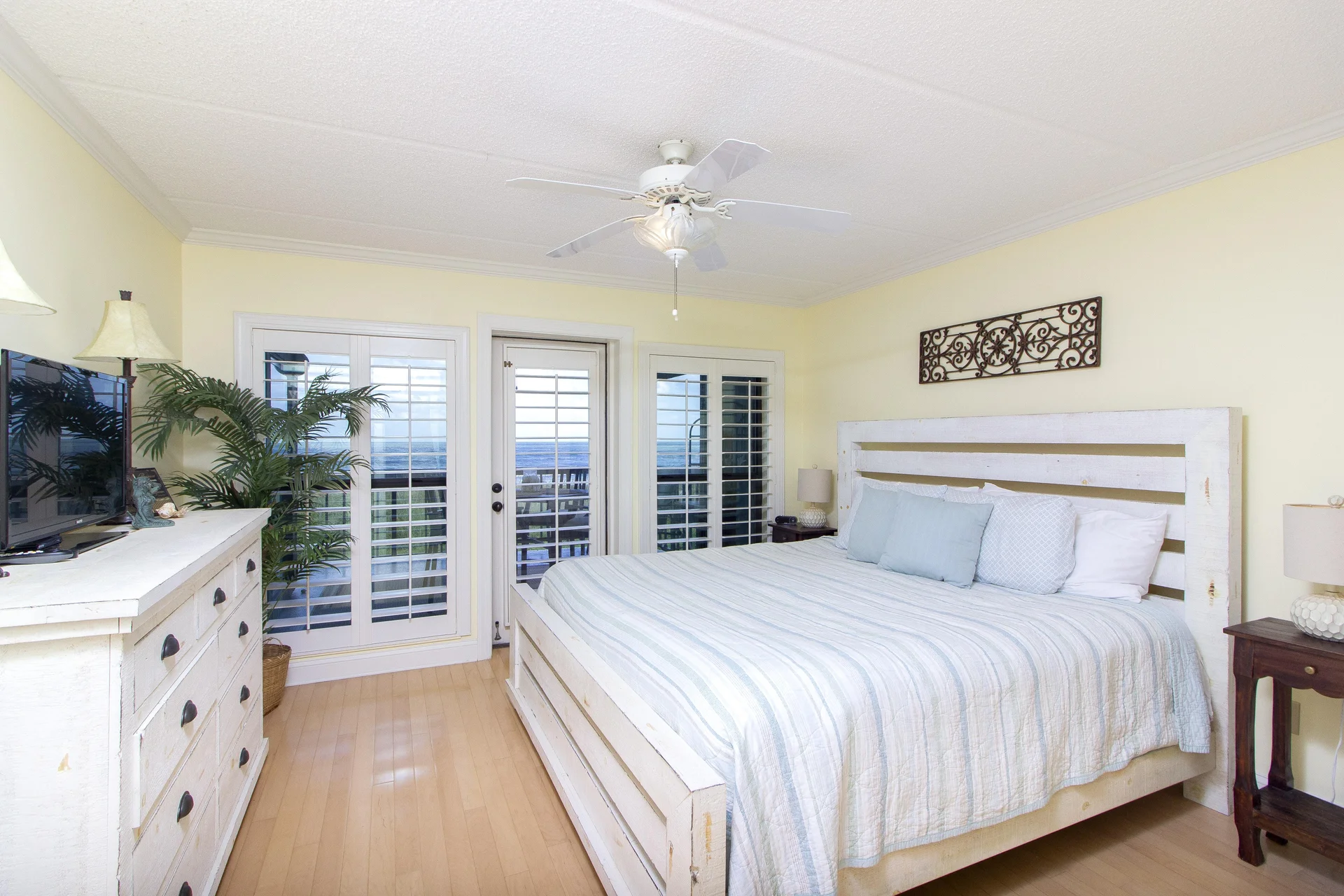 Primary bedroom with whitewashed wood bed frame, striped bedding, plantation shutters, and ocean view balcony doors.
