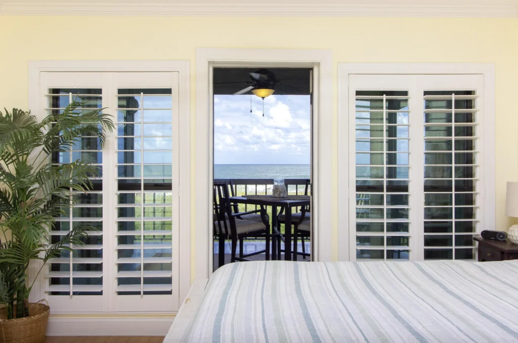 Striped bedding covers the bed as plantation shutters frame a glass door opening onto the oceanfront balcony.