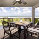 Covered balcony dining area with dark wood table, cushioned chairs, ceiling fan, and ocean views beyond railing.