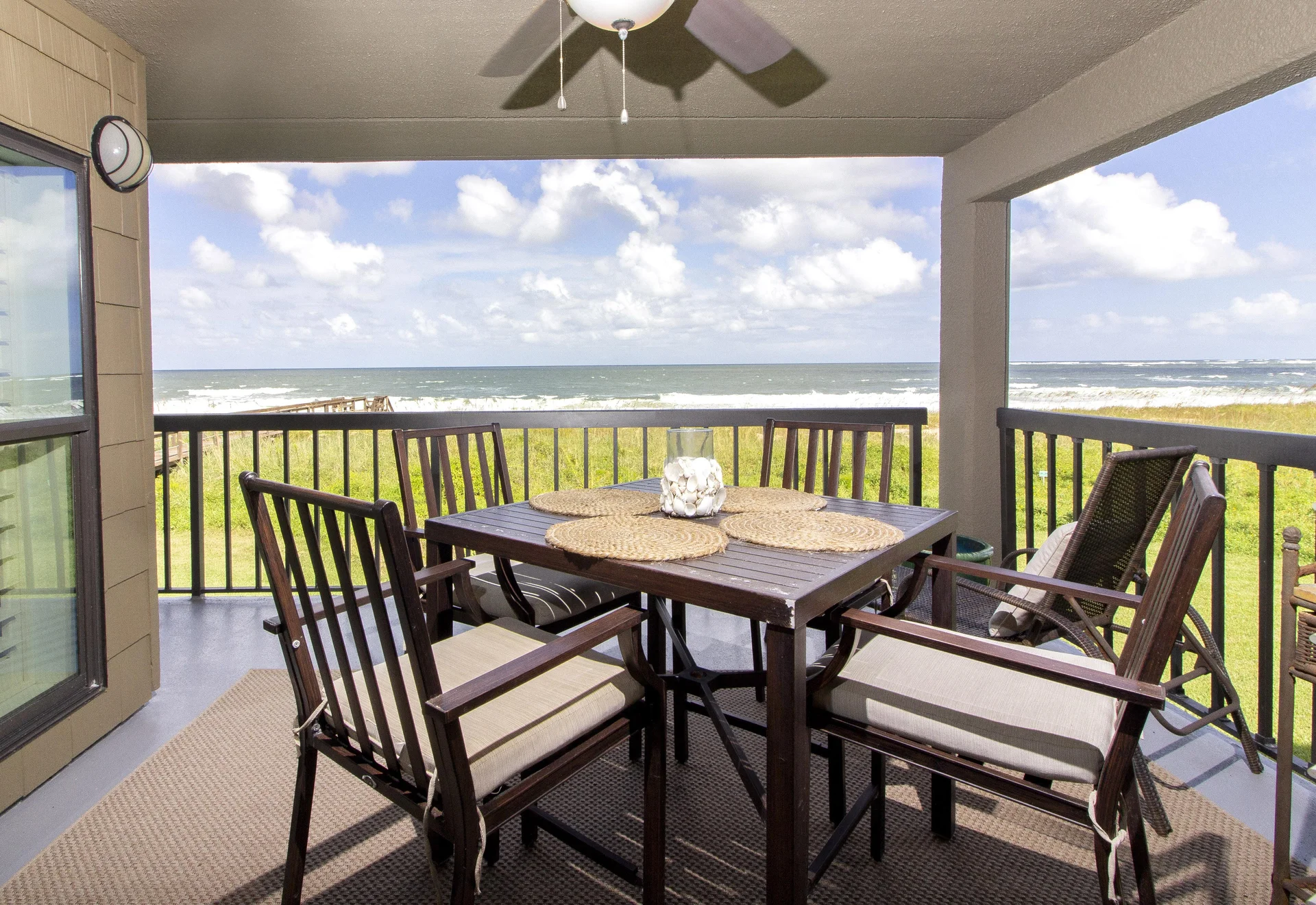 Covered balcony dining area with dark wood table, cushioned chairs, ceiling fan, and ocean views beyond railing.