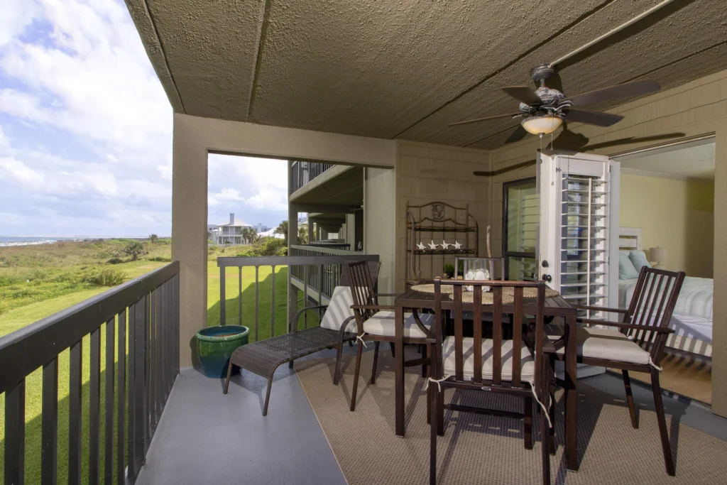 Second-floor balcony with cushioned dark wood chairs, woven chaise lounge, ceiling fan, and grassland views.