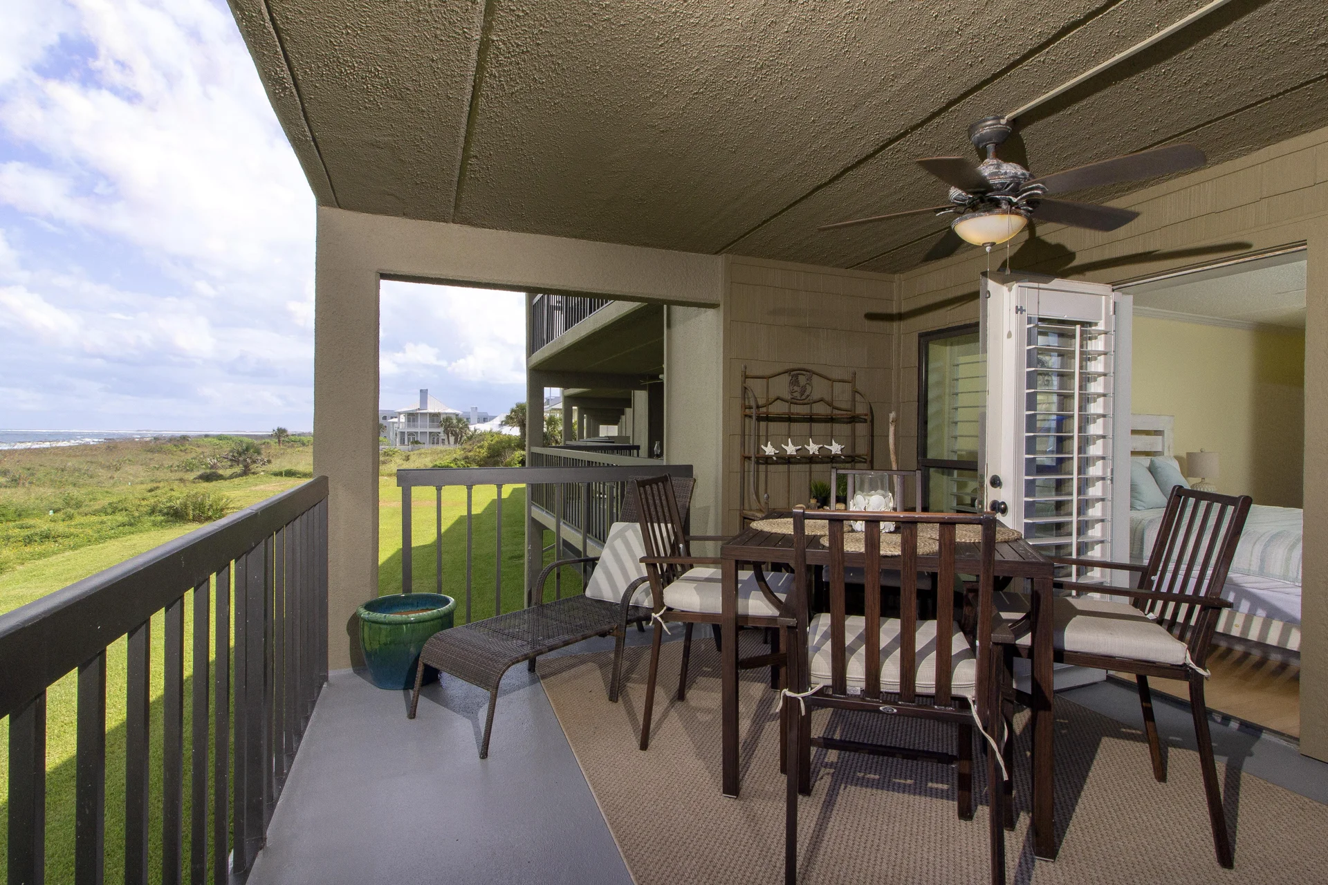Second-floor balcony with cushioned dark wood chairs, woven chaise lounge, ceiling fan, and grassland views.