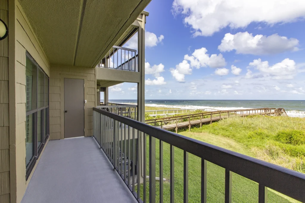 Second-floor balcony with gray concrete floor, metal railing, beige siding, and ocean views over grassy dunes.