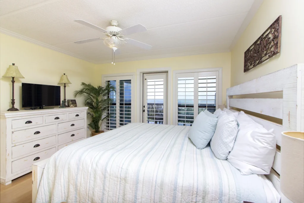 King bed with striped bedding and multiple pillows faces plantation-shuttered doors opening to an ocean-view balcony.