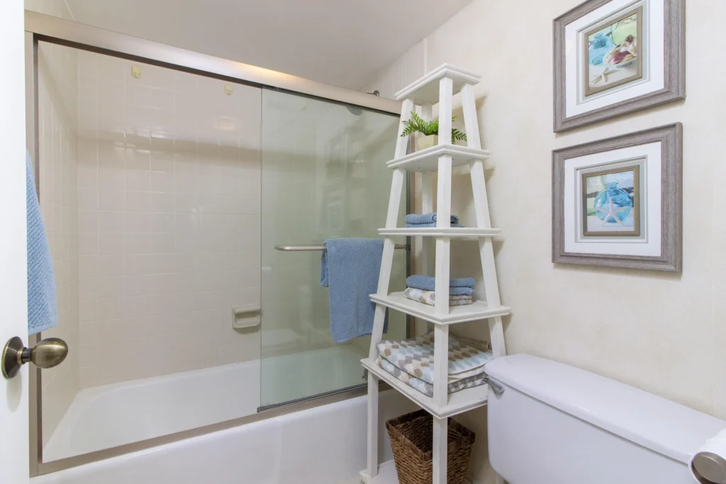 Bathroom with white tiled tub and sliding glass doors, white tiered shelf holding towels and decor.