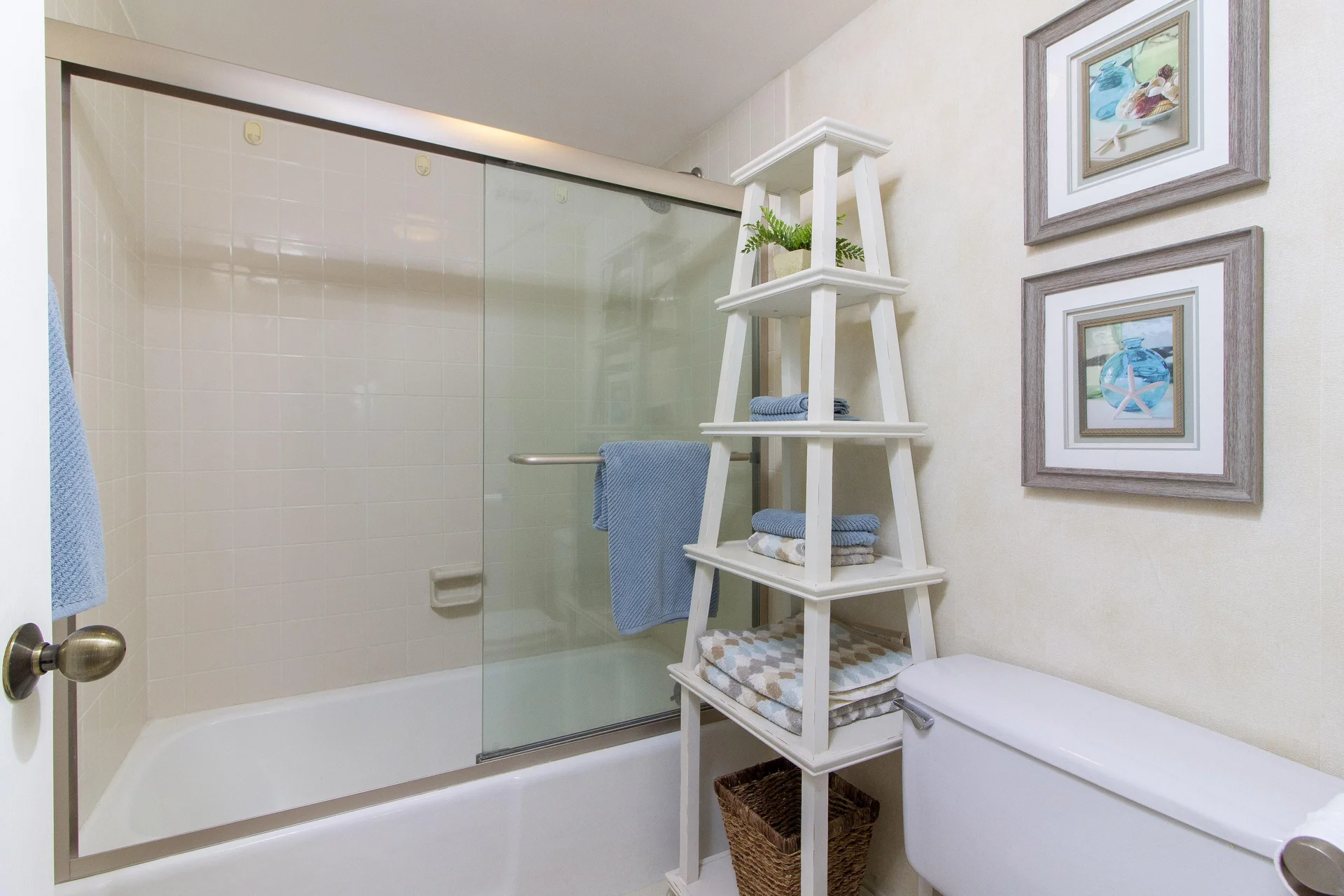Bathroom with white tiled tub and sliding glass doors, white tiered shelf holding towels and decor.
