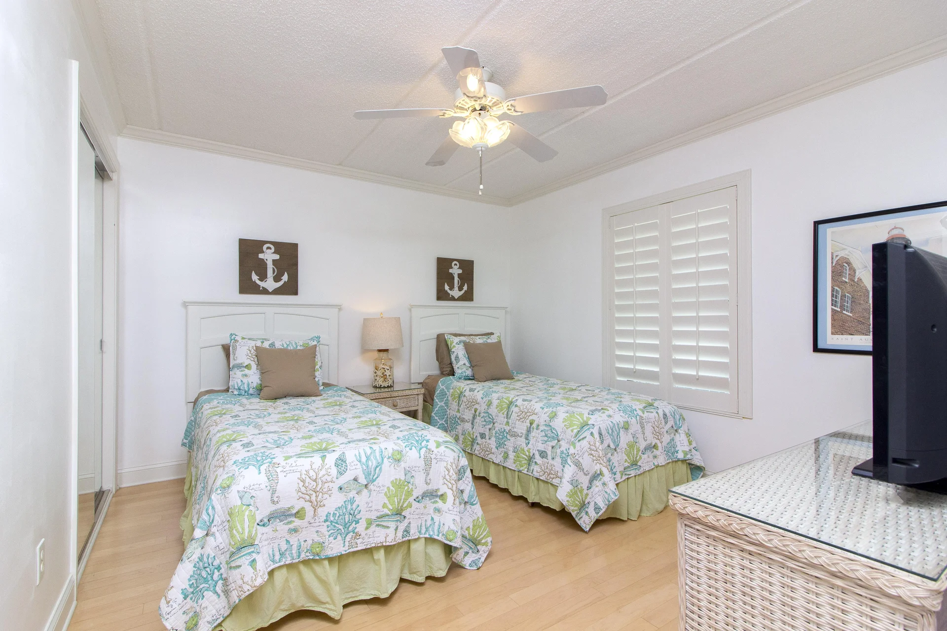 Bedroom with twin beds dressed in seashell-patterned quilts, wicker nightstand, ceiling fan, and plantation shutters.