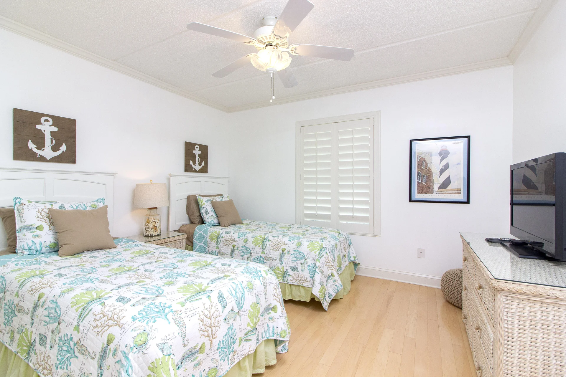 Bedroom with twin beds dressed in sea life quilts, white headboards, wicker dresser, and shuttered window.